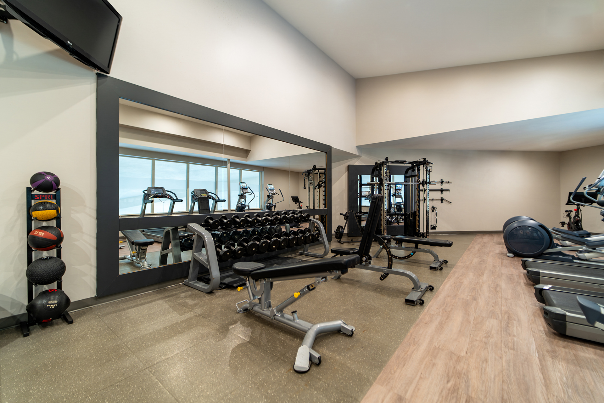 A large fitness center with light wood accents, a large mirror, cardio equipment, free weights and benches.