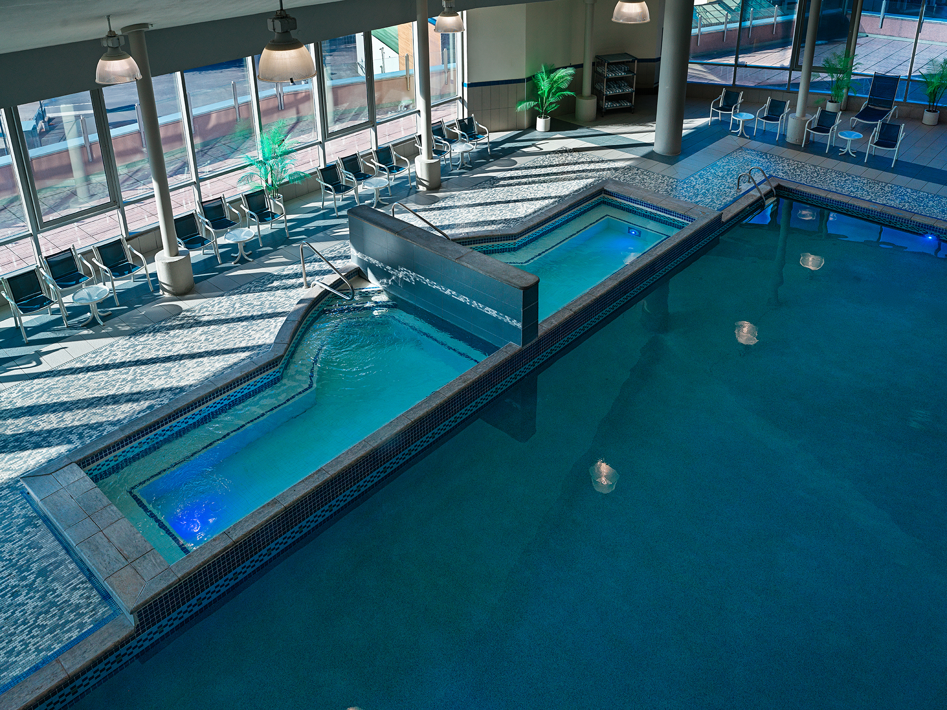 A large indoor swimming pool, with two hot tubs connected to the pool, large windows surrounding the pool deck with tables and lounge chairs.