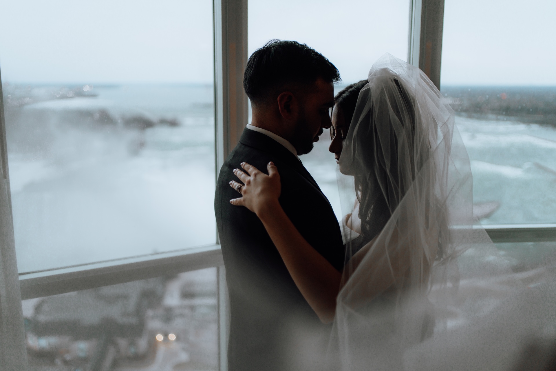 A bride and groom embracing in front of a large window overlooking Niagara Falls.