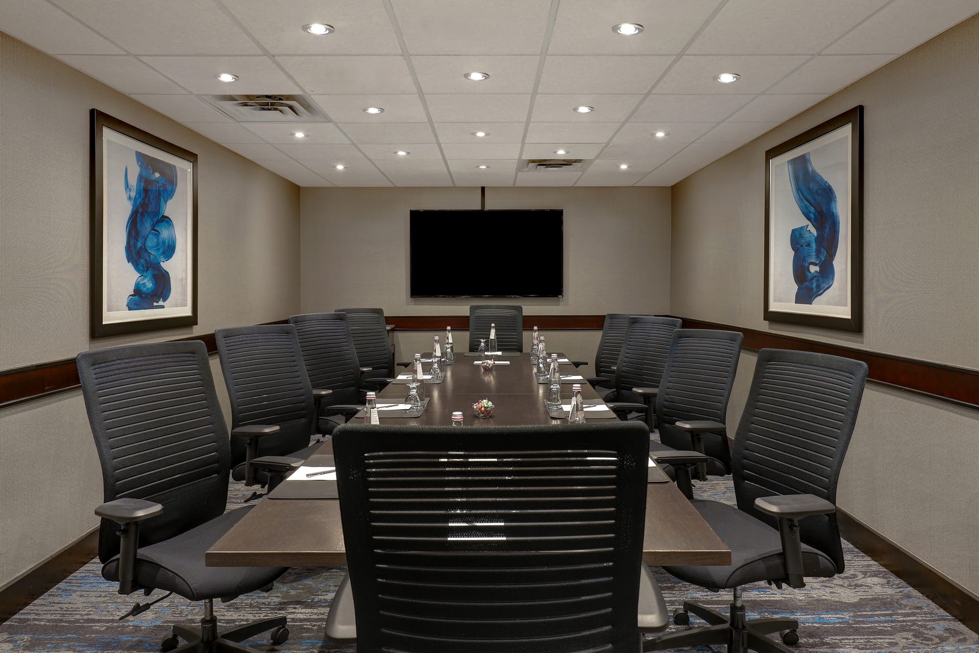 A meeting room with a large conference table surrounded by large chairs, state-of-the-art built in audio/visual components and a large display screen.