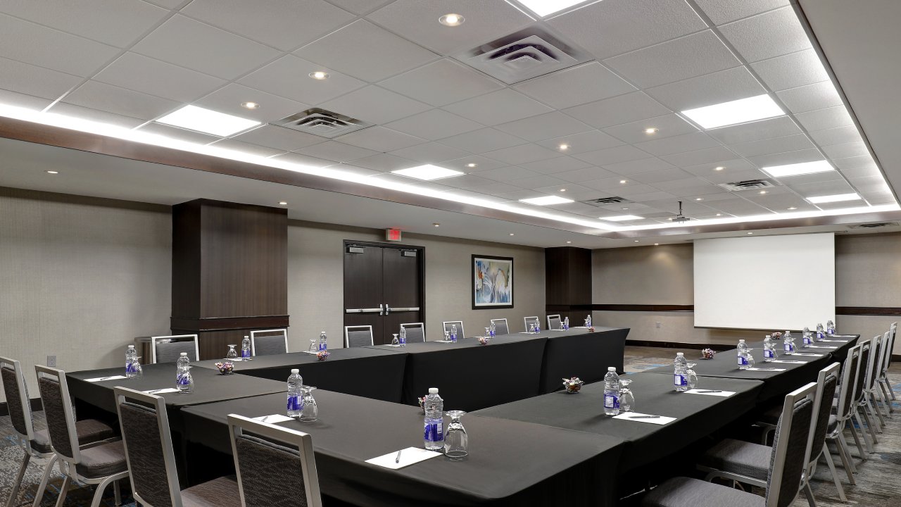 Large, bright meeting room with spacious seating and large able in u-shape facing a large projector screen.