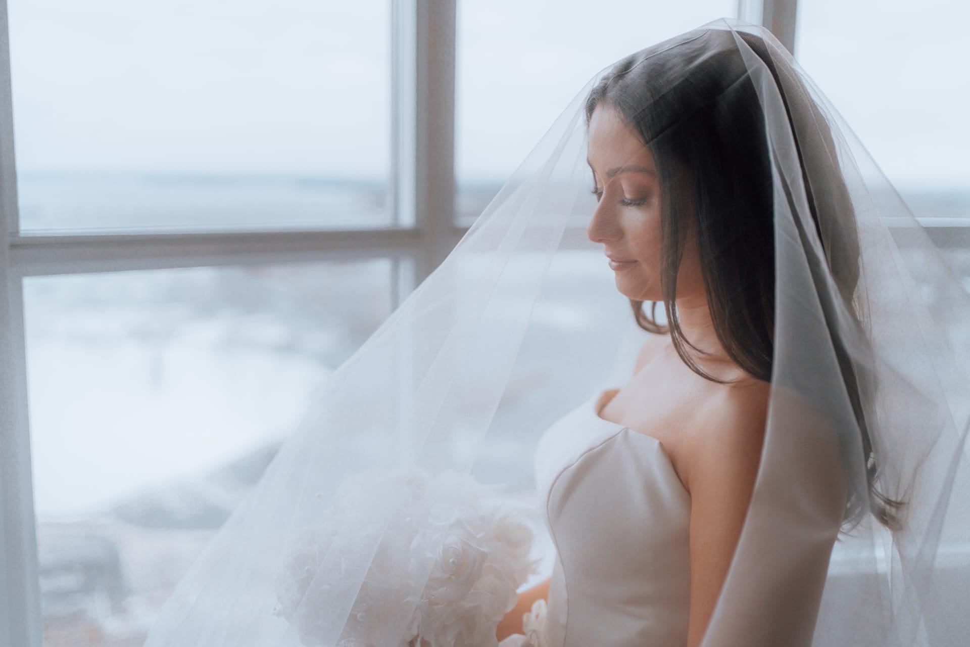 A bride wearing a veil looking down at a white bouquet of roses standing in front of a large window overlooking Niagara Falls.